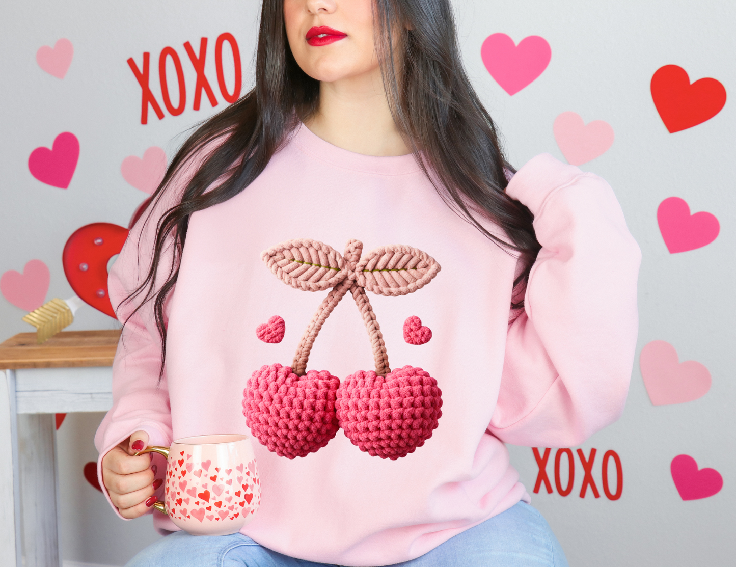 Person wearing a pink sweatshirt with cherry design, holding a mug, surrounded by heart and 'XOXO' decorations.