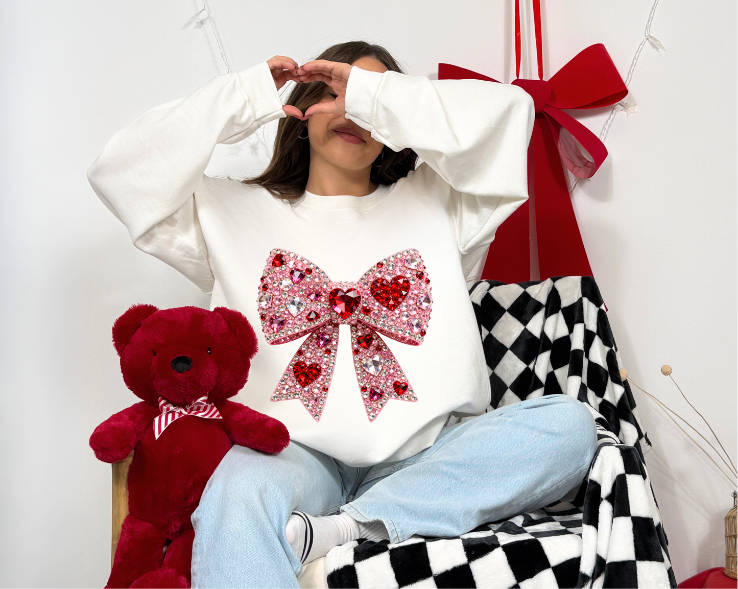 Person wearing a white sweatshirt with a large red bow design, sitting next to a red teddy bear and a checkered chair.