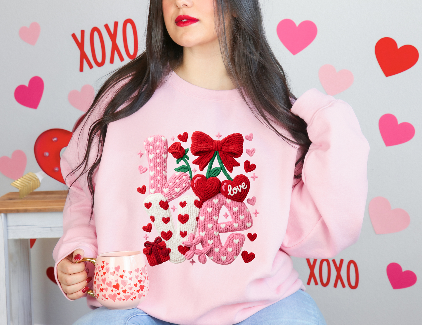 Person wearing a pink sweatshirt with Valentine's Day designs, holding a mug, with decorative hearts and 'XOXO' text in the background.