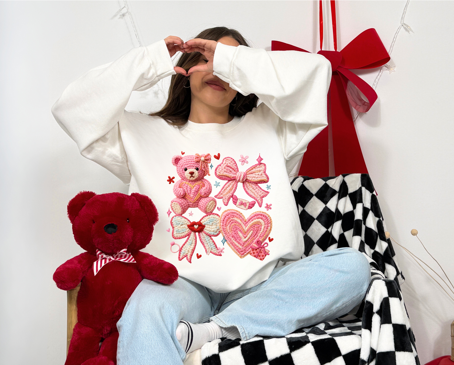 Person wearing a white sweatshirt with pink designs, sitting next to a red teddy bear on a checkered floor.